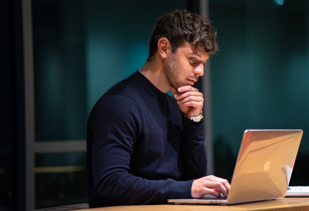 Man in a dark sweater sitting at a laptop with chin resting on hand in deep concentration, representing distraction-free focused work